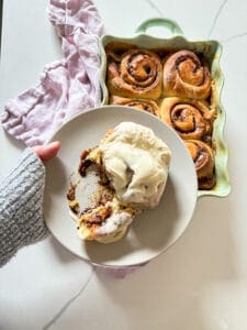 Hand with a sweather holding a plate with a frosted cinnamon roll and a pan of cinnamon rolls