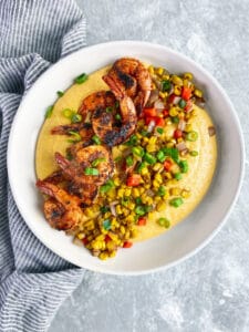 white bowl with yellow cheese grits, blackened shrimp, and sautéd onions, peppers and corn.