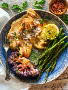 Dover Sole with white wine sauce on a blue plate with purple cauliflower and asparagus and a lemon