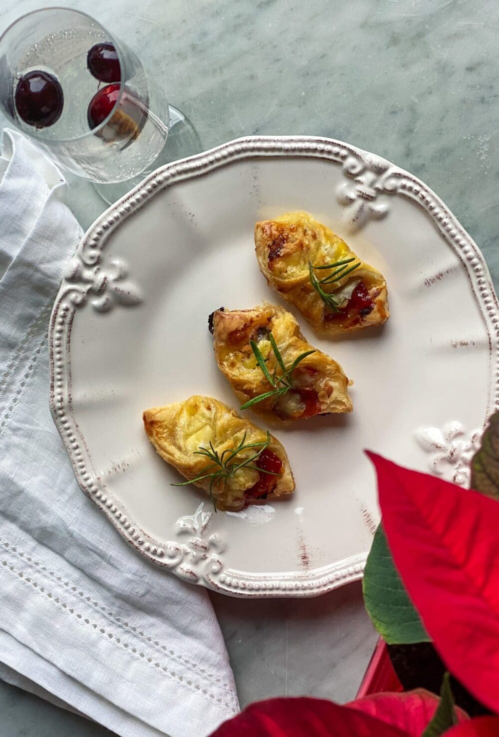 3 puff pastry wrapped brie, prosciutto, and pepper jelly bites, with a glass of champagne, with cranberries, and a poinsettia