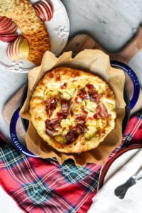 Baked brie with prosciutto, pepper jelly and rosemary in a pan with pears, apples and crackers next to a plaid napkin