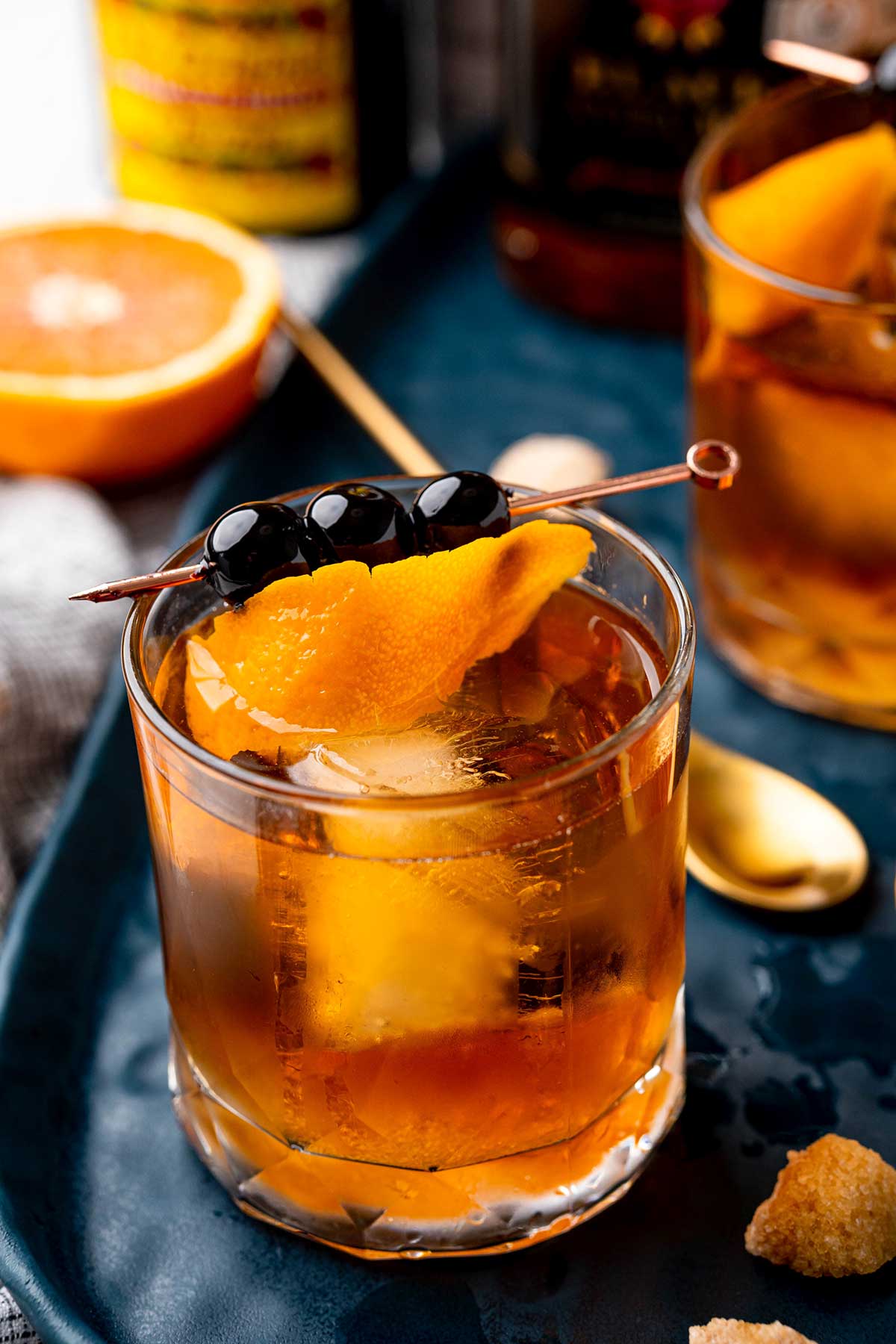 Old fashioned cocktail with cherries and an orange peel with a spoon and orange half next to it on a tray
