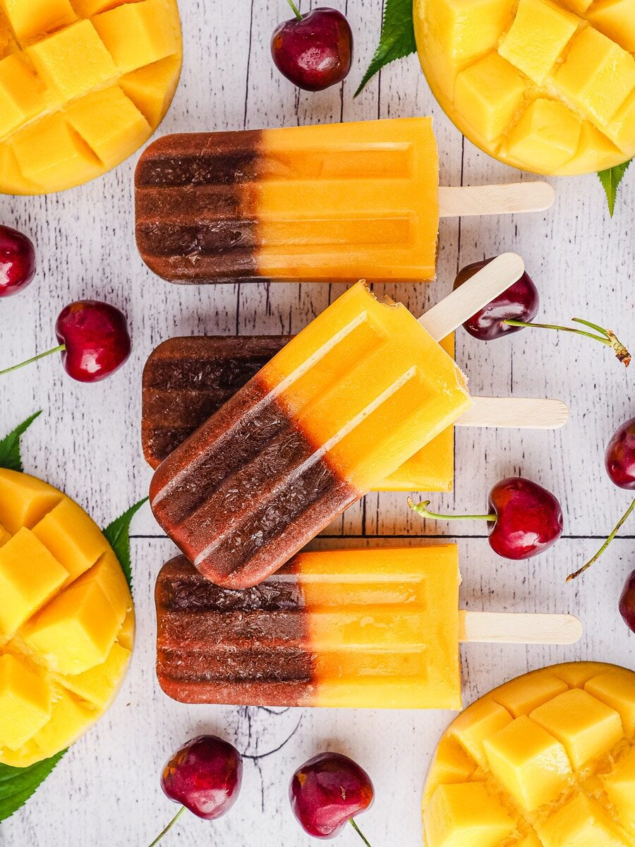 mango cherry popsicles laying on a whitewashed table with mango and cherries
