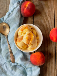 peach icecream in a bowl with a silver spoon on a bue stripe napkin with 3 peaches