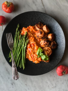 Tomato risotto with shrimp and asparagus on a black plate with a fork and tomatoes on the side