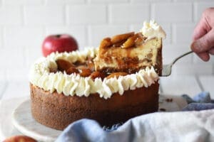 Slice of caramel apple cheesecake being taken out of the entire cheesecake and red apples and blue napkin