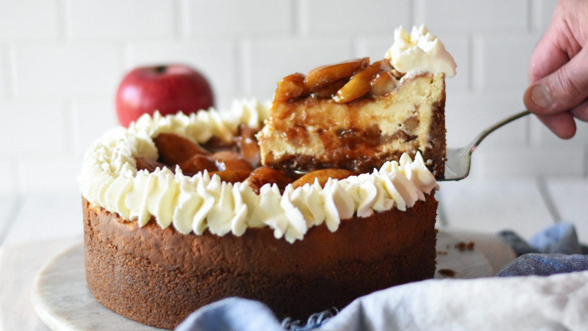 Slice of caramel apple cheesecake being taken out of the entire cheesecake and red apples and blue napkin