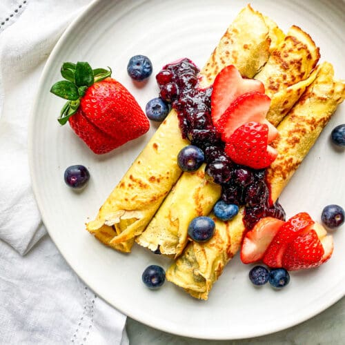 crepes rolled up on a white plate with blueberry compote and sliced strawberries