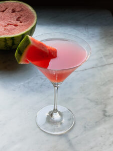 watermelon martini in a martini glass with a wedge of watermelon and a half of watermelon in background
