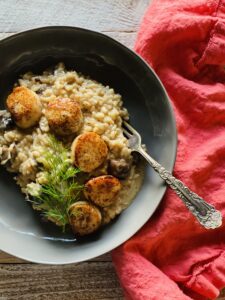 Bowl of mushroom risotto with scallops and dill on top and a bright orange napkin