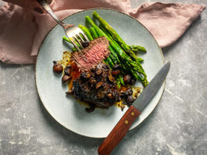 Medium rare steak piece on a fork with a plate with mushrooms and asparagus