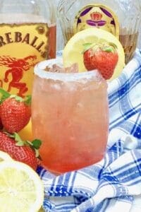 Strawberry Spiked Lemonade with Fireball Whisky garnished with a lemon and a strawbery with a blue and white napkin and a bottle of fireball