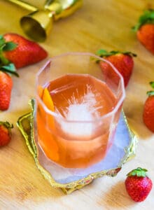 a strawberry old fashioned in a glasss with ice and orange peel garnish on a coaster. Strawberries laying on table