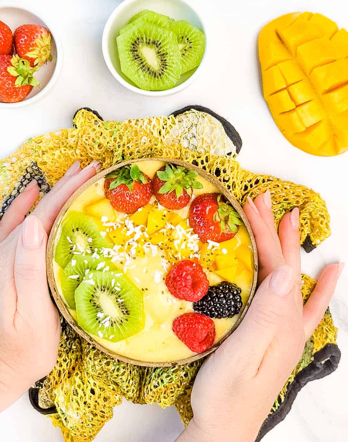 two hands holding a mango smoothie bowl with kiwi, rasperries, blackberries and strawberies on top