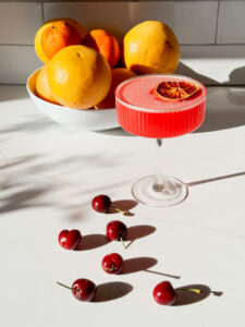 Cherry sea breeze martini with 3 cherries skewered on top, a bowl of graperfruits oranges and a few cherries