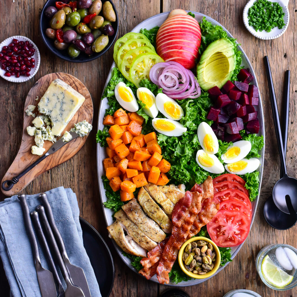 cob slad ingredients on a platter with blue cheese and olives in a bowl and blue napkins and utensils