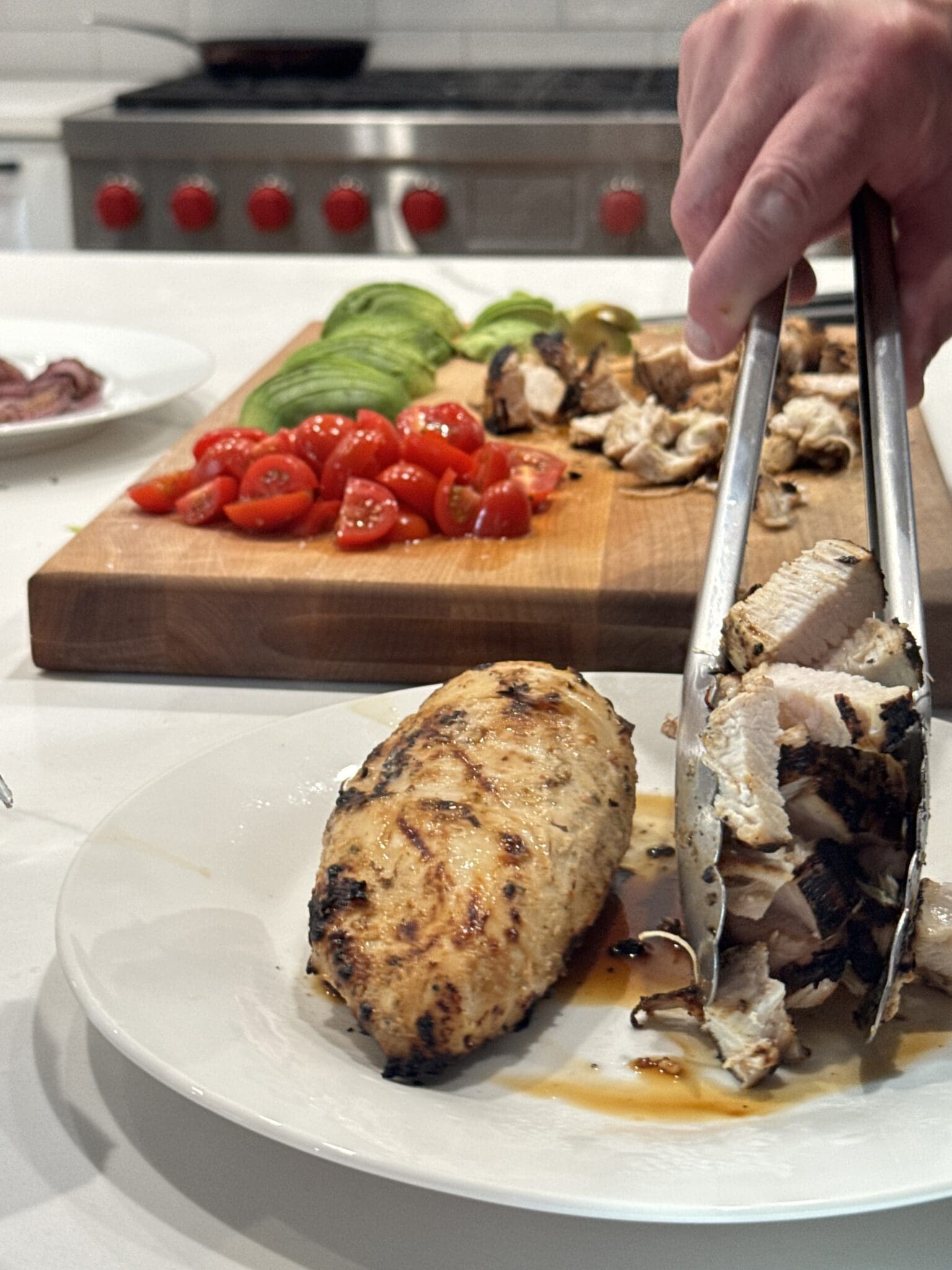 grilled chicken being held by tongs and a cutting board with tomatoes and avocadoes