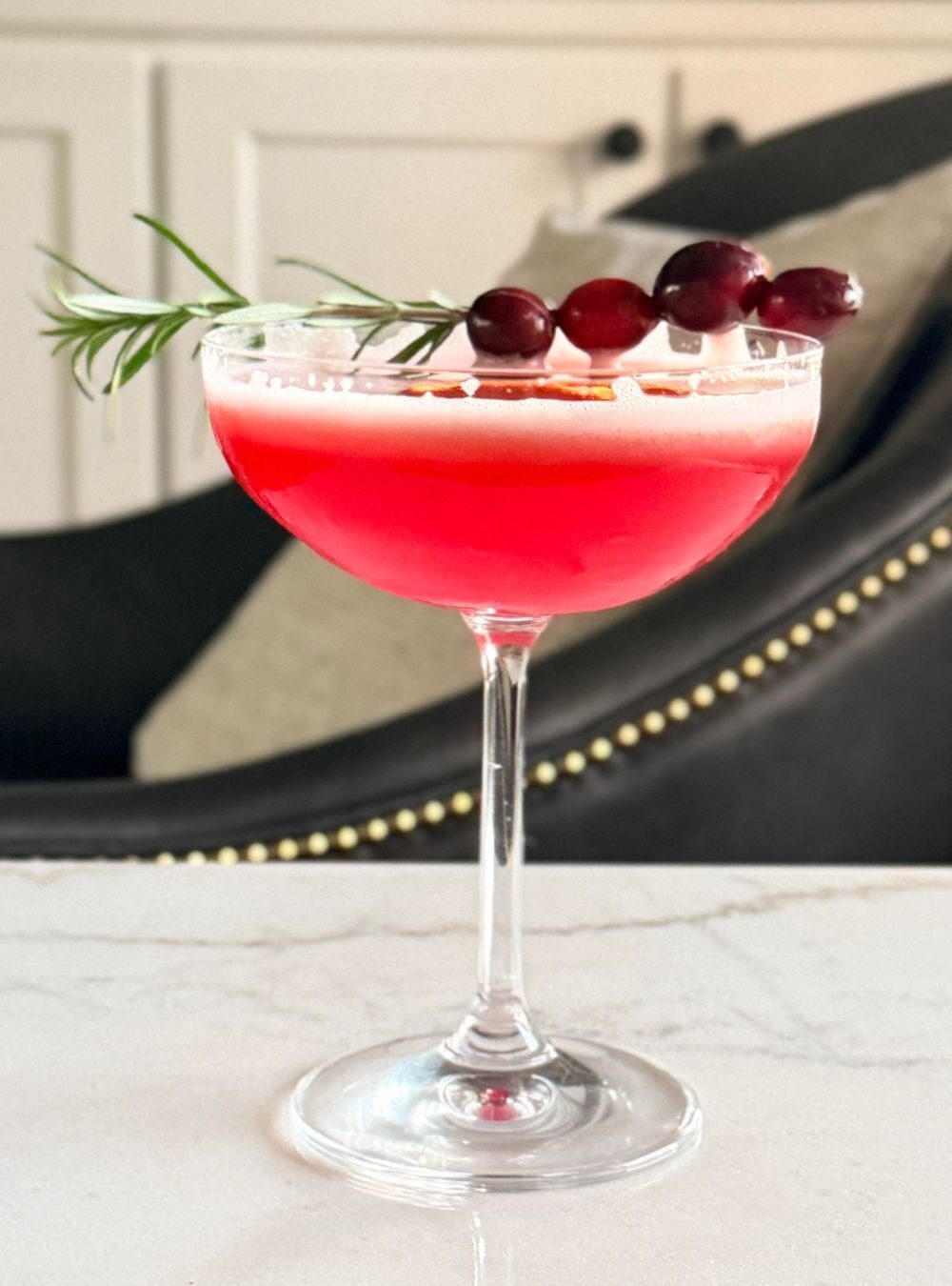 Cranberry orange whiskey sour in a coupe glass with cranberries on rosemary sprig from the side.