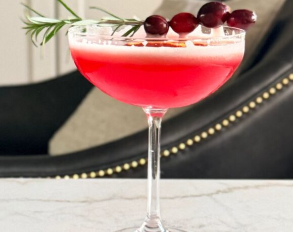Cranberry orange whiskey sour in a coupe glass with cranberries on rosemary sprig from the side.