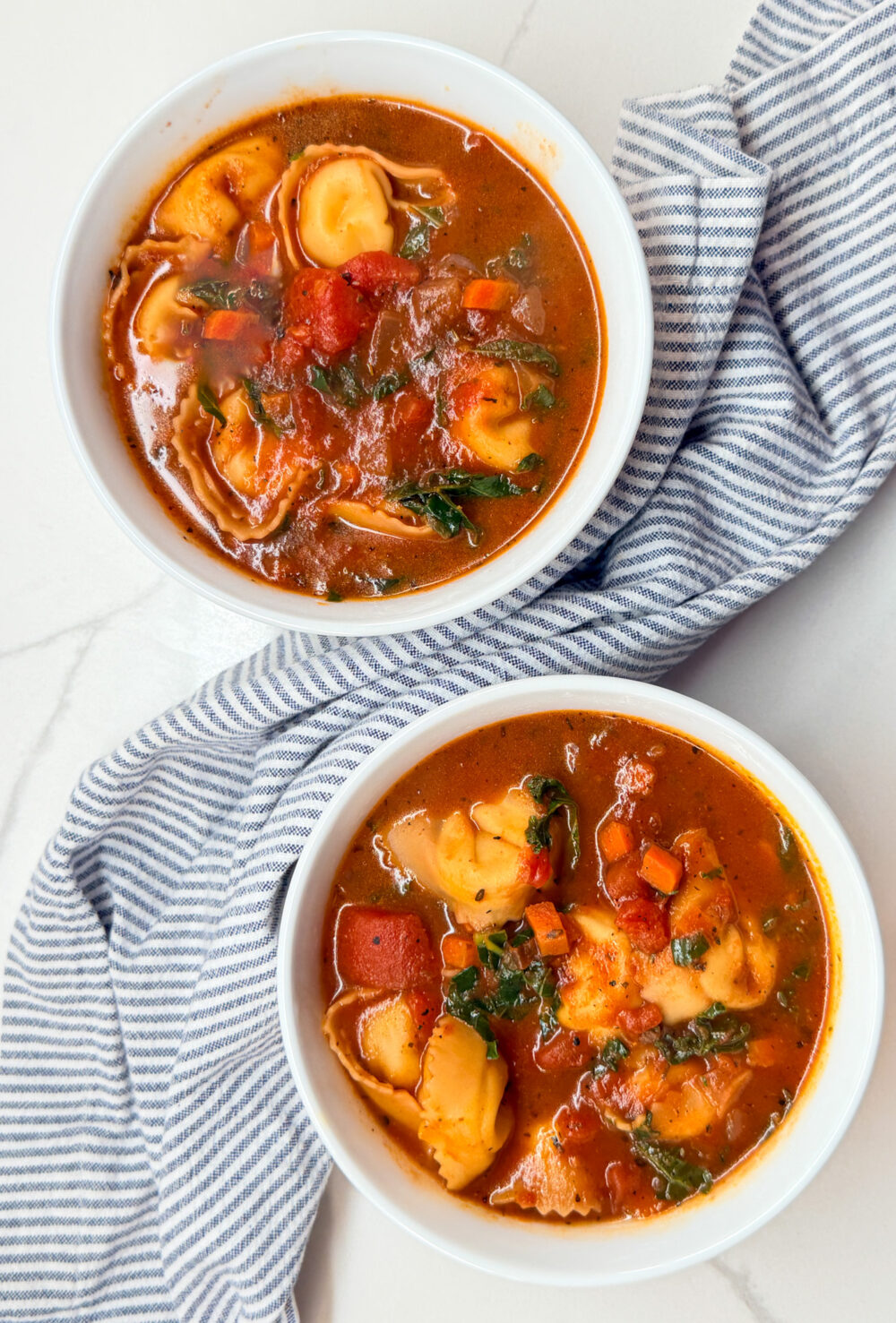 2 bowls of tortellini tomato and kale soup with a blue and white napkin
