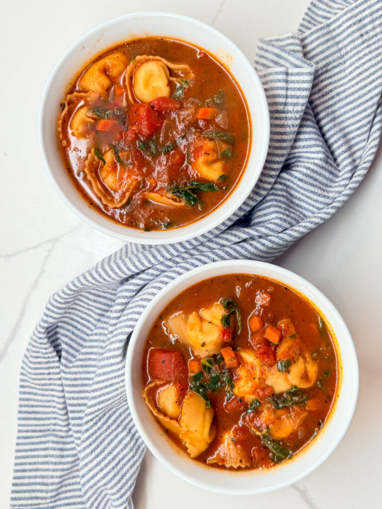 2 bowls of tortellini tomato and kale soup with a blue and white napkin