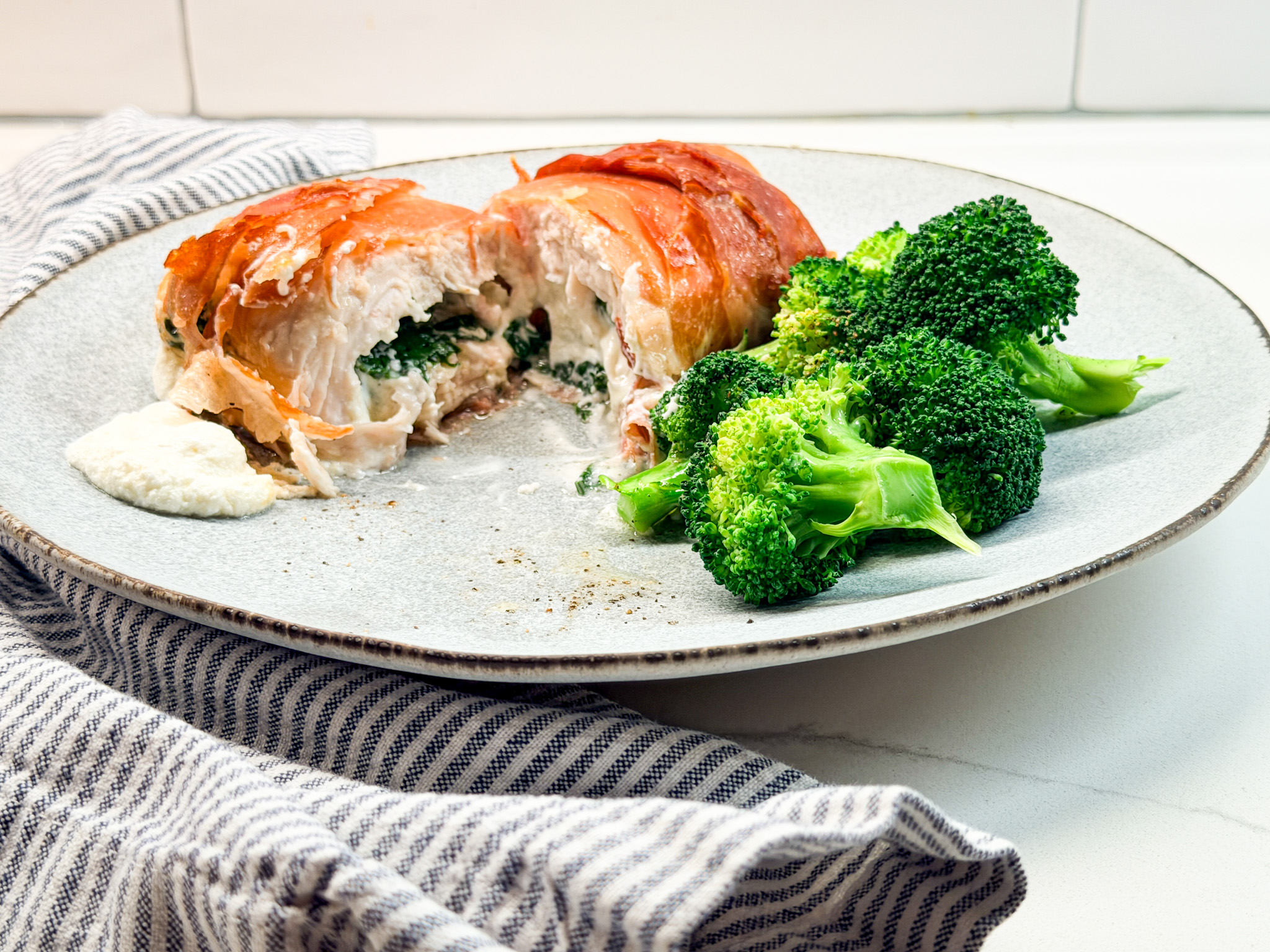 Proscuiotto wrapped chicken filled with goat cheese and spinach on a plate with broccolli