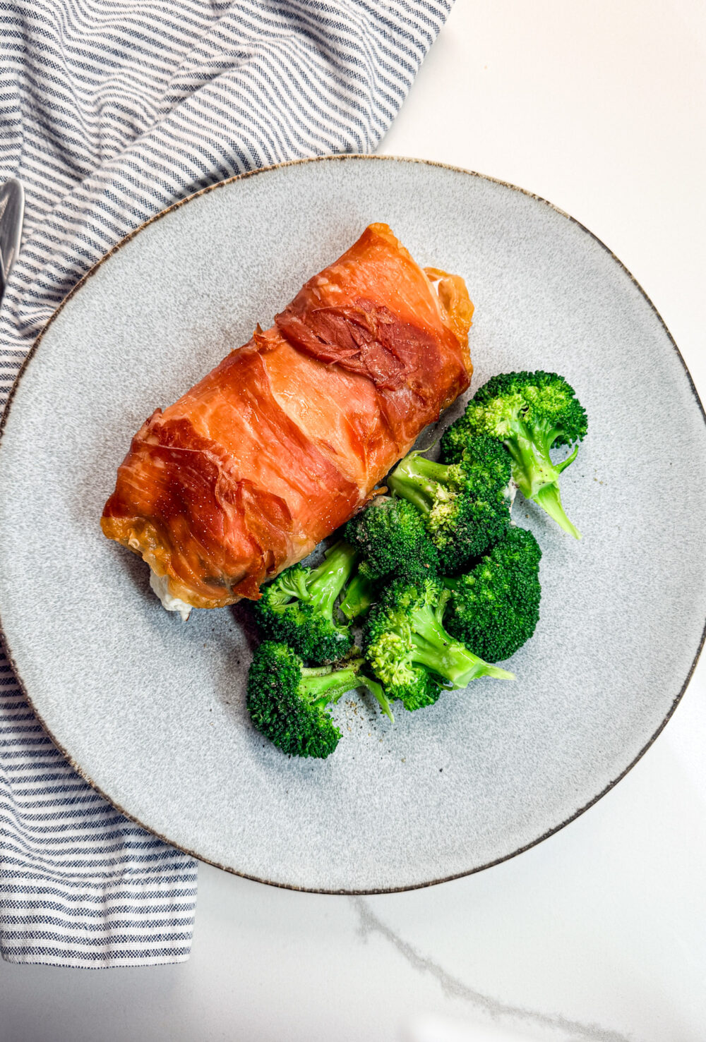 proscuiotto-wrapped chicken on a plate with broccolli and a napkin and fork
