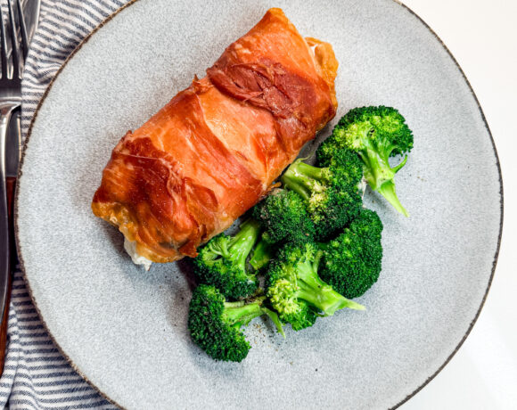 proscuiotto-wrapped chicken on a plate with broccolli and a napkin and fork