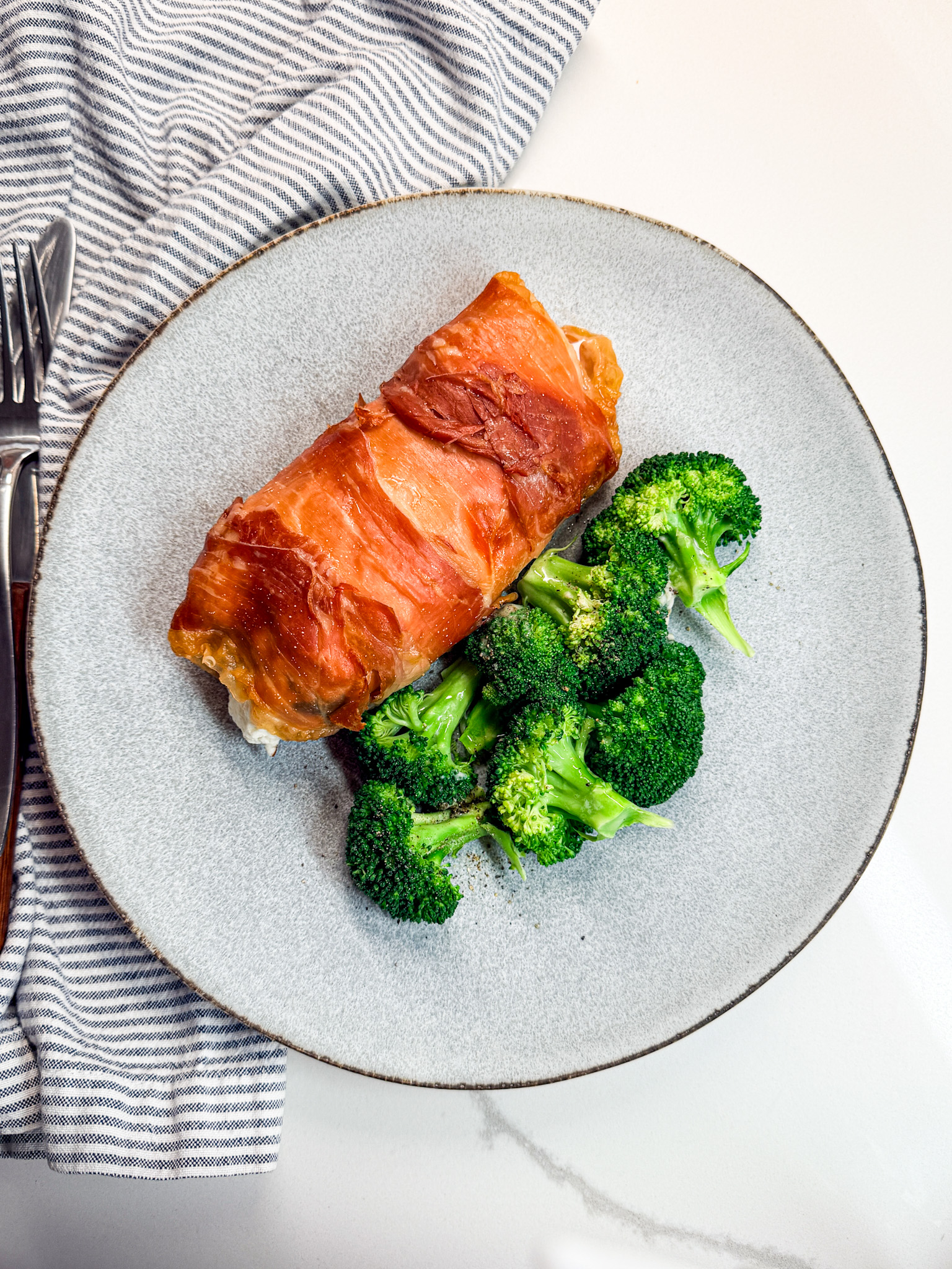 proscuiotto-wrapped chicken on a plate with broccolli and a napkin and fork